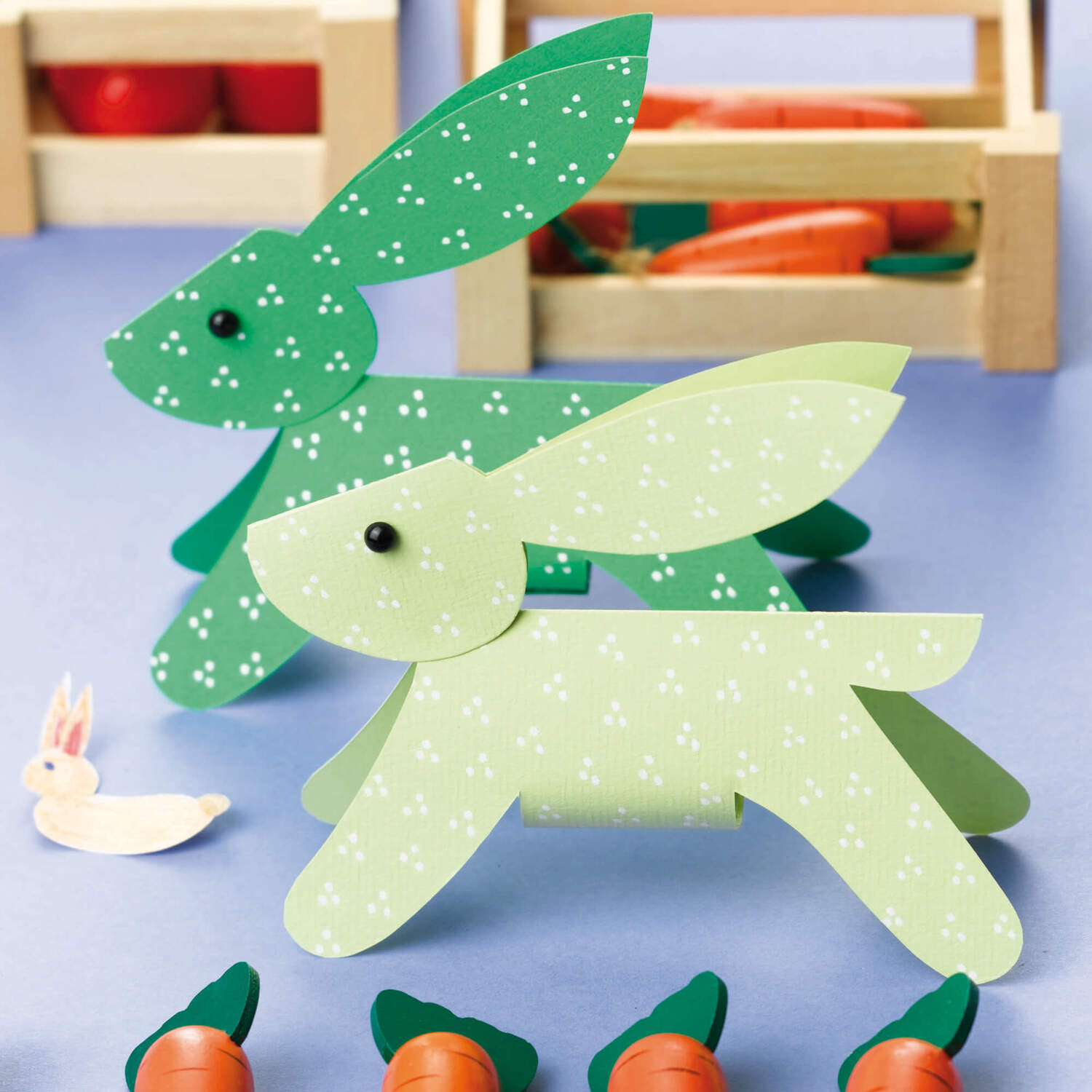 Bastelanleitung Osterhase aus Papier: Zwei Papierhasen in Grün und Hellgrün mit Karotten im Hintergrund.