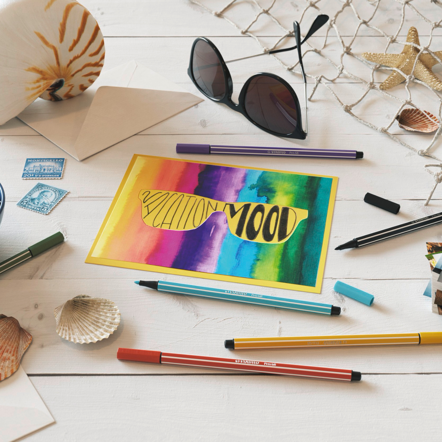 Pen 68 auf einem weißen Tisch mit Muscheln und Postkarten. Bunte Stifte und Sonnenbrille ergänzen das kreative Arrangement.