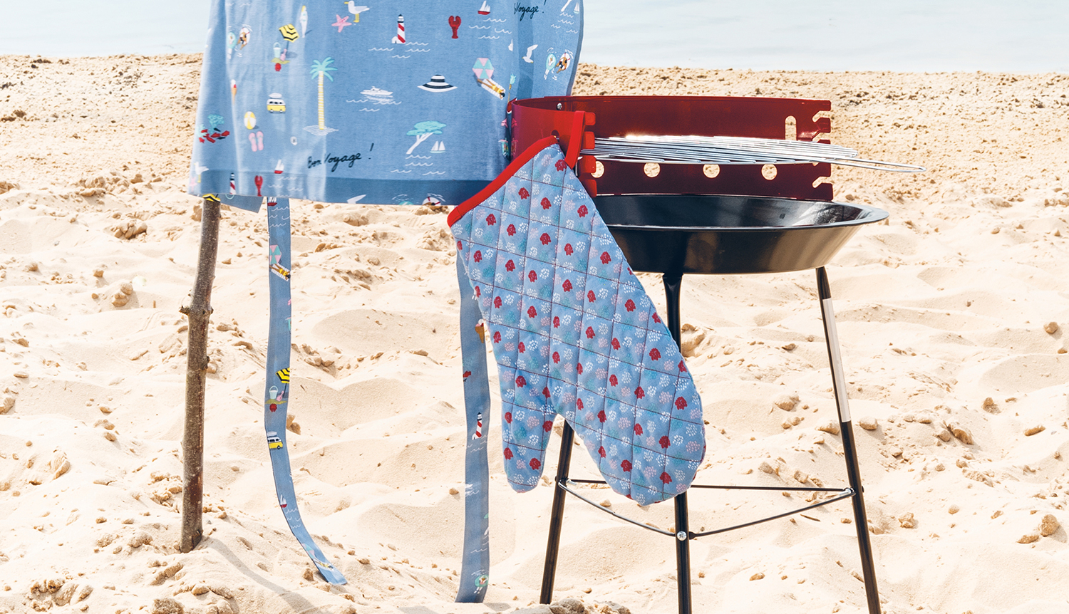 Nähanleitung Grillhandschuh: Ein blauer Grillhandschuh mit maritimem Muster hängt an einem Grill am Strand.