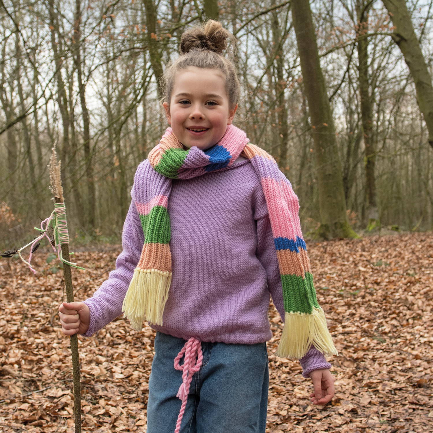 Strickanleitung Schal aus Essentials Soft Merino aran: Kind im Wald mit buntem Schal und lila Pullover.
