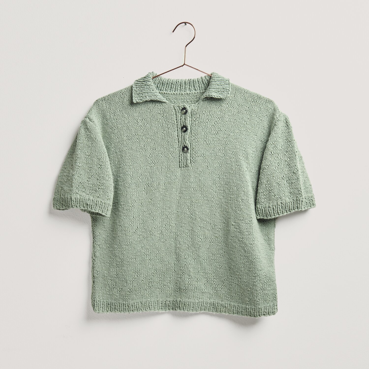 Strickset Shirt Modell 03a: Kurzarmshirt in Salbei, gestrickt aus Essentials Super Cotton dk, präsentiert auf einem Kleiderbügel.