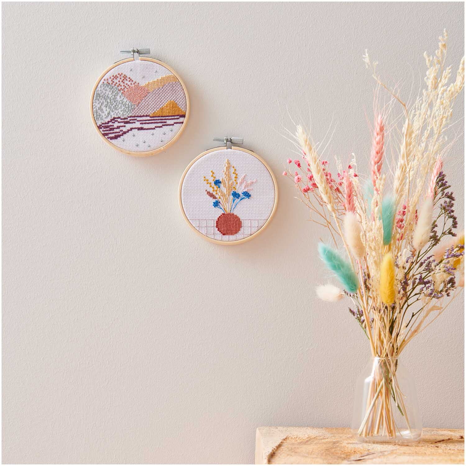 Stickpackung Mini Kit Trockenblumen Bild Ø 10cm zeigt bunte Stickereien an einer Wand. Daneben steht ein Strauß Trockenblumen in einer Vase.