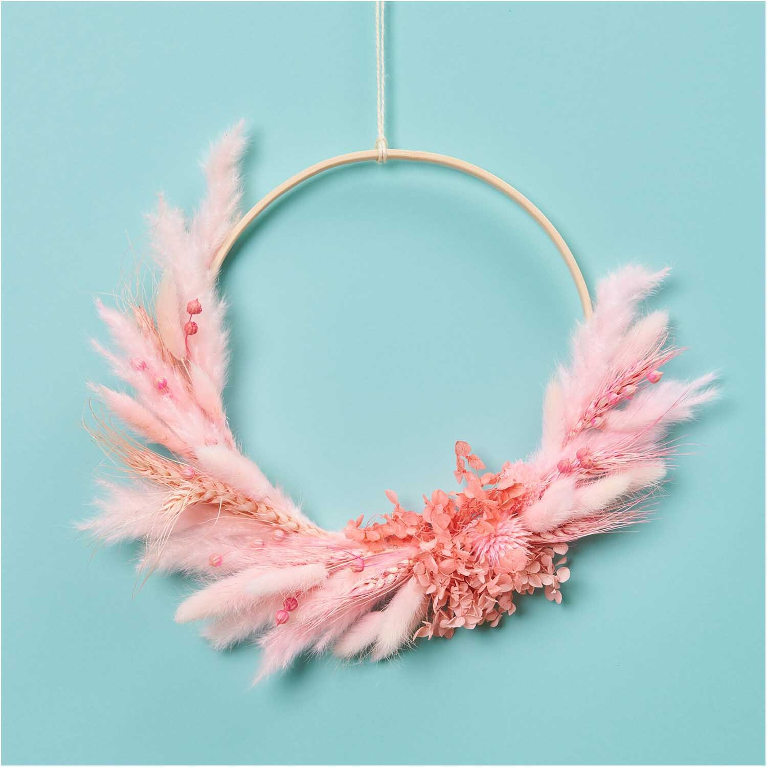 Getrockneter Weizen pink 60-70cm 50 Stück: Ein dekorativer Kranz aus rosa getrocknetem Weizen von Rico Design vor blauem Hintergrund.