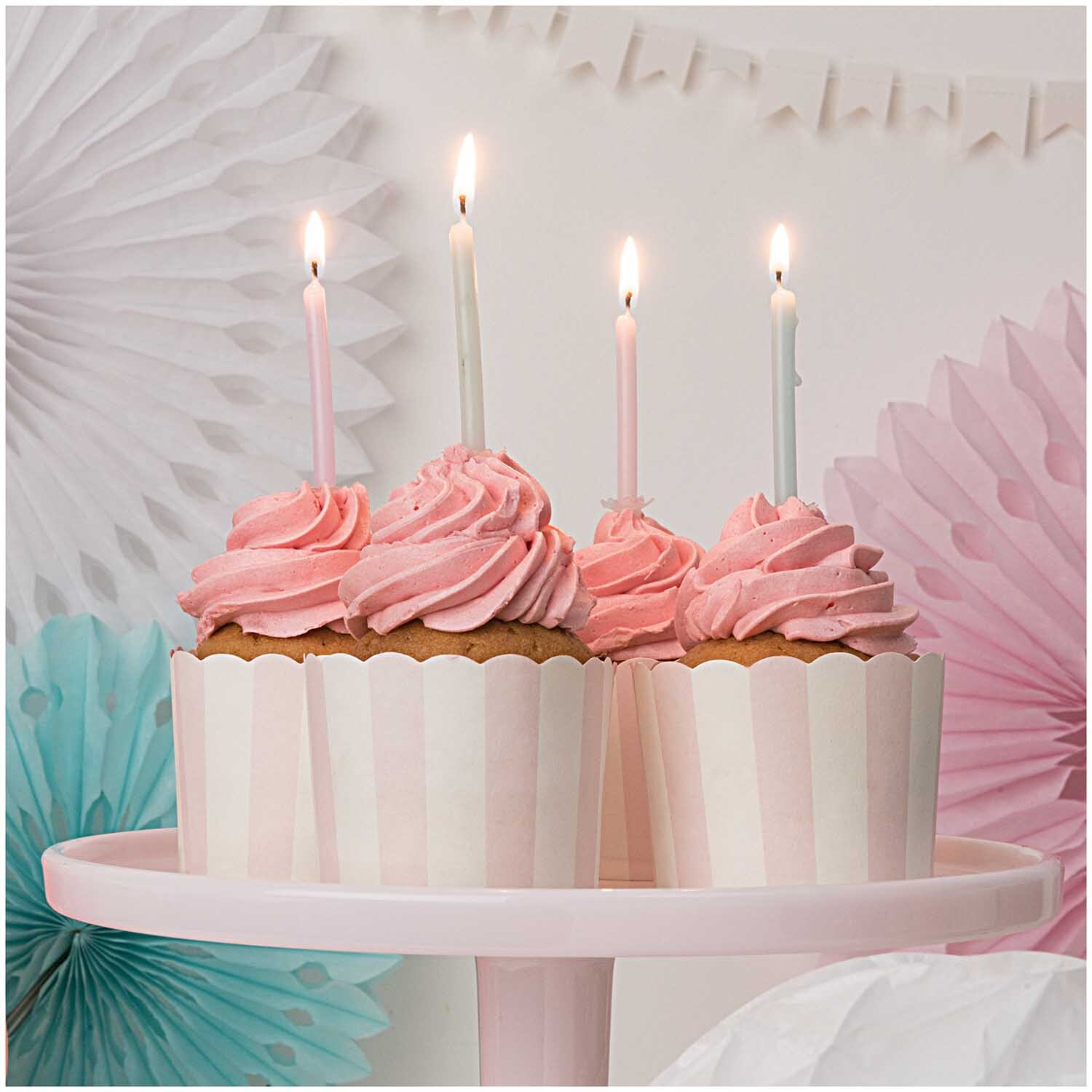 Wabenpapier Blumen Sommer 3 Stück: Vier rosa Cupcakes mit Kerzen auf einem rosa Teller, umgeben von dekorativen Papierblumen.