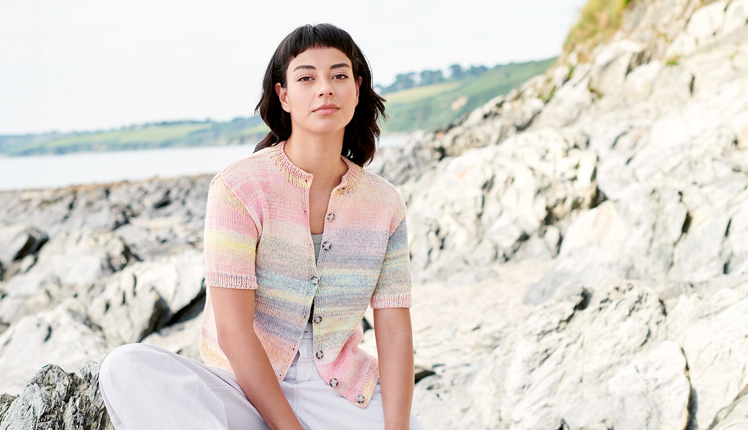 Strickanleitung Jacke Creative Chic-Unique Cotton dk, sommerliche Kurzarmstrickjacke in Pastelltönen, lässig getragen von Frau auf Felsenküste am Meer
