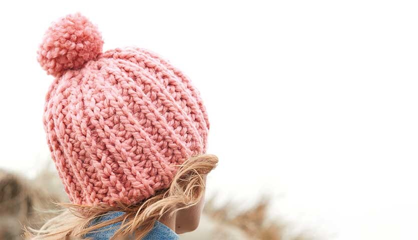 Strickanleitung gerippte Mütze aus Creative Twist super chunky: Rosa Mütze mit Bommel, ideal für kalte Tage, getragen von einer Person im Freien.