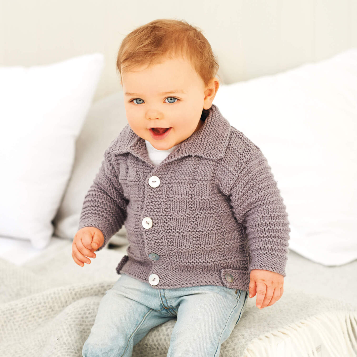 Strickanleitung Babyjacke mit Kragen: Ein Baby trägt eine graue Strickjacke mit Kragen und weißen Knöpfen, sitzt auf einem Bett.