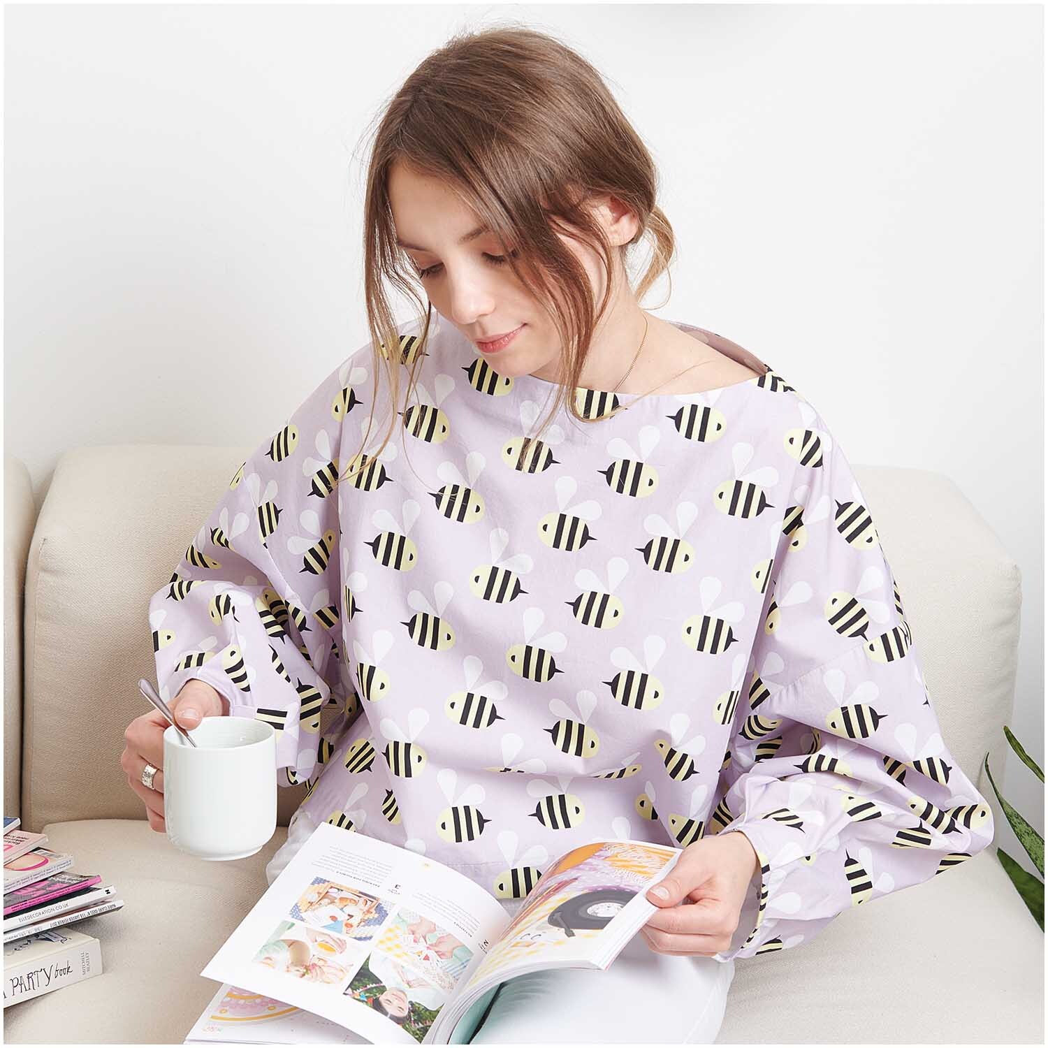 Stoffabschnitt Baumwoll-Popelin flieder Bienen: Frau in fliederfarbenem Oberteil mit Bienenmuster, liest ein Buch und hält eine Tasse.