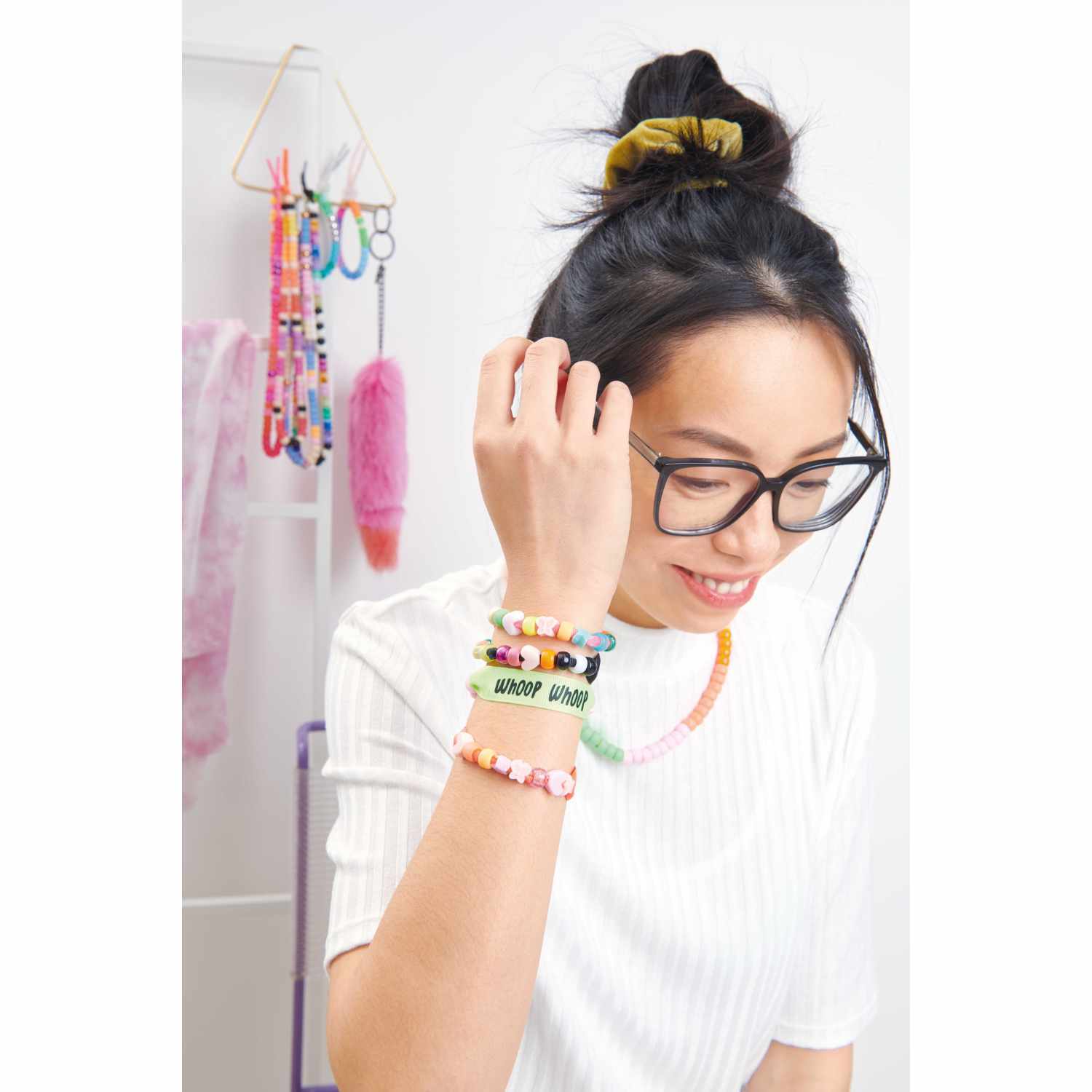 Itoshii - Ponii Beads Bügelmotiv Schriftzüge 8 Designs: Frau mit bunten Armbändern und Brille, lächelnd in einem kreativen Raum.