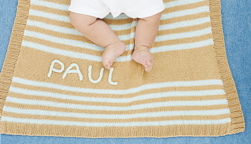 Strickanleitung Babydecke mit Streifen: Beige-weiße Decke mit Namen, Babyfüße auf blauem Hintergrund.