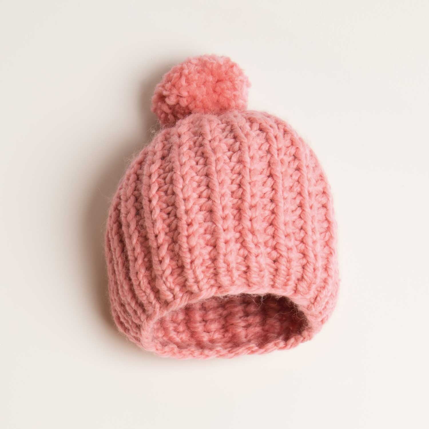 Strickanleitung gerippte Mütze aus Creative Twist super chunky: Rosa Mütze mit Bommel, gestrickt in Rippenmuster, liegt auf hellem Hintergrund.