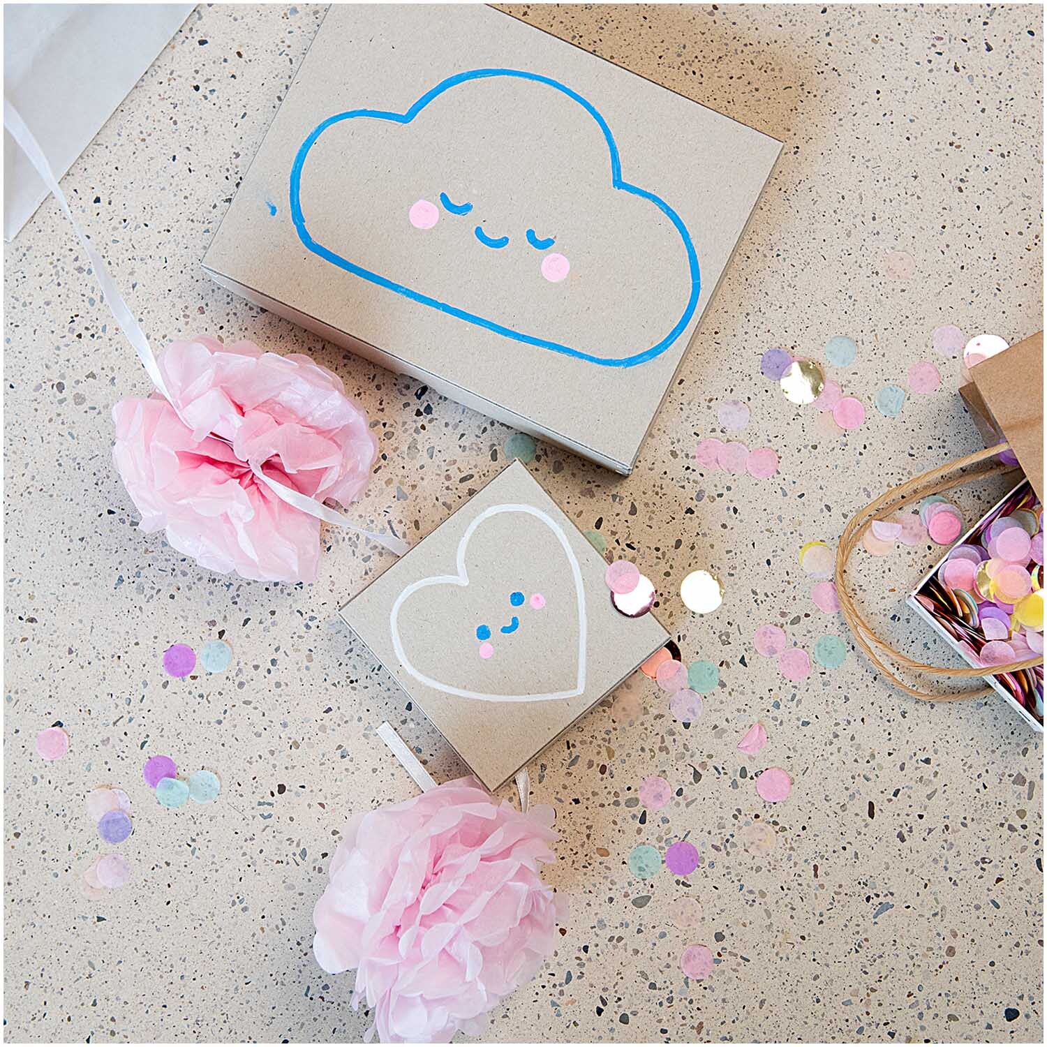 Kreidestifte Flüssigkreide Set Regenbogen: Kreative Geschenkverpackungen mit Wolken- und Herzmotiven, dekoriert mit buntem Konfetti und rosa Blumen.