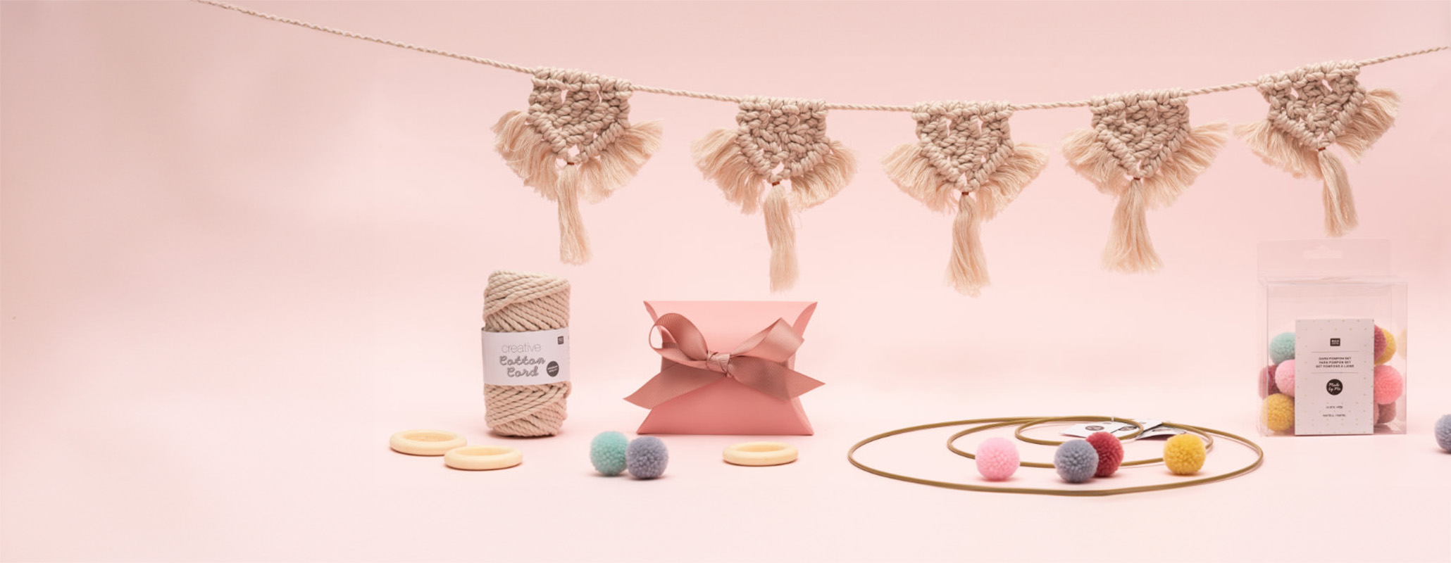 Hängende Makramee-Girlande mit Garn, eine Geschenkschachtel, verschiedene Dekoringe und Verpackung mit Garn-Pompons. Fotosetting rosa Wand.