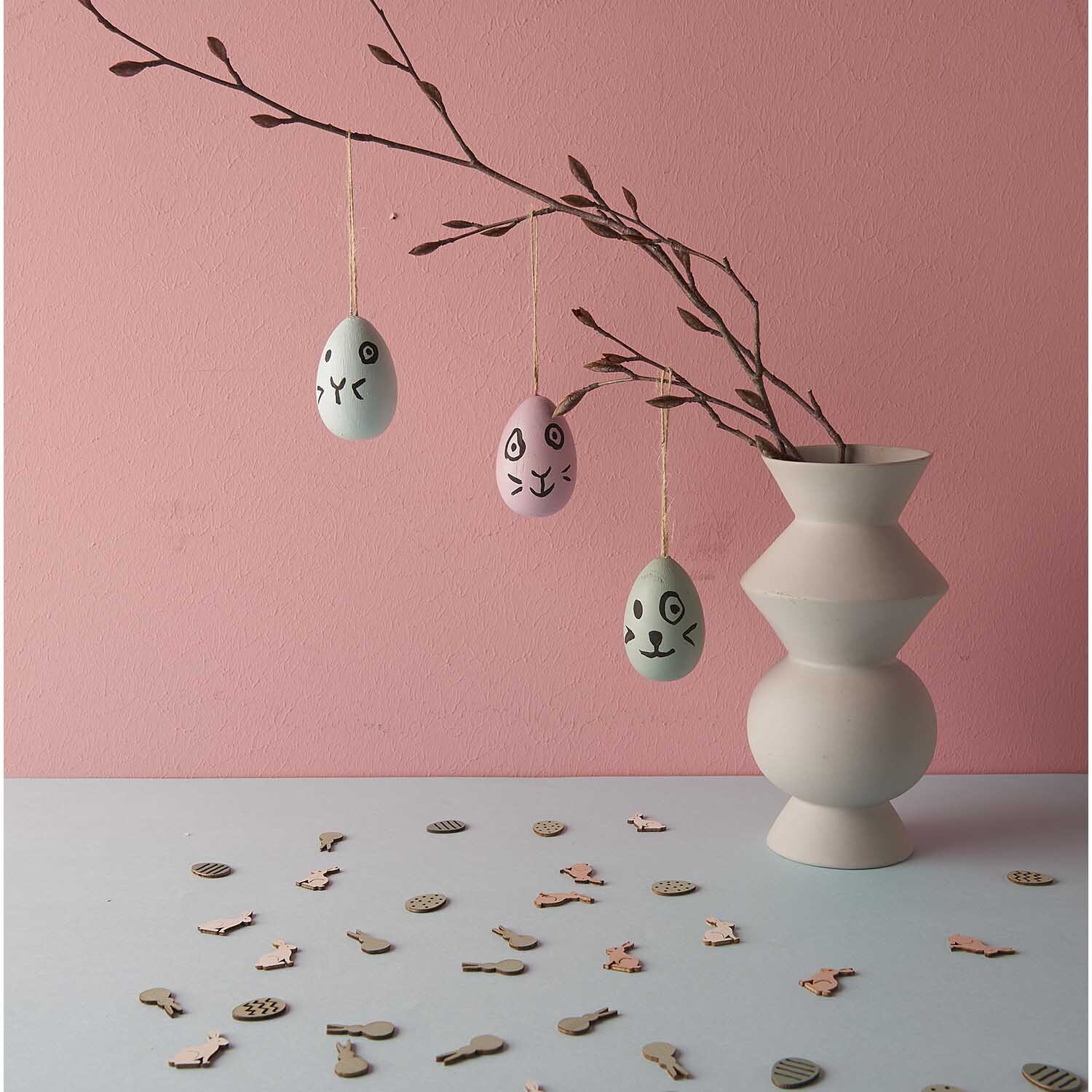 Holzeier zum Hängen 5 Stück: Drei dekorative Eier an Zweigen in einer modernen Vase vor rosa Wand.