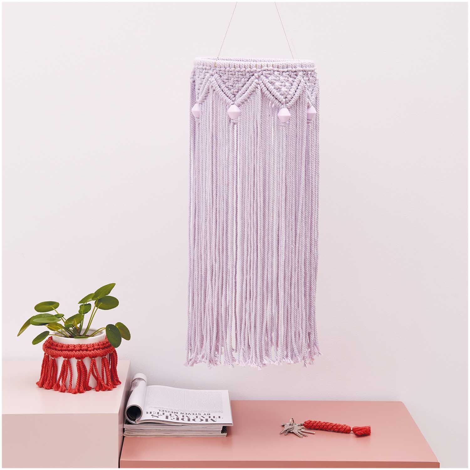 Makramee Set Mobilé aus Rico Macrame: Lila Wandbehang mit langen Fransen, dekorativ über einem Tisch mit Pflanze und Notizbuch.