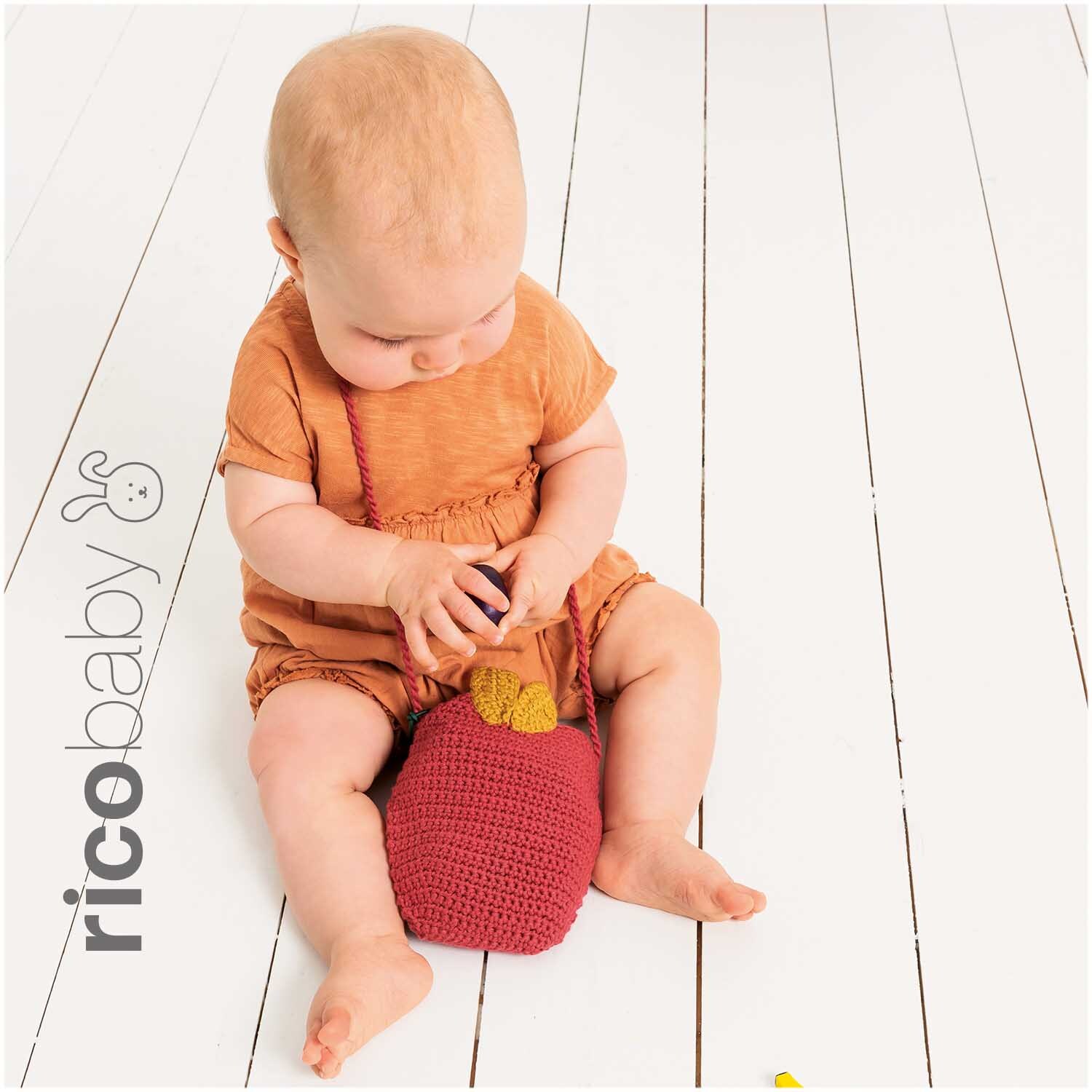 Rico Baby Cotton Soft dk: Ein Baby in orangefarbenem Outfit spielt mit einer himbeerfarbenen Tasche.