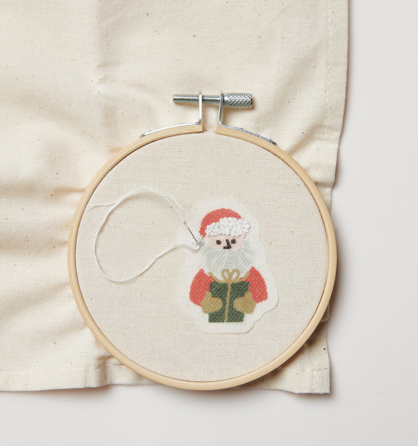 Stick & Stitch Stickpackung Weihnachtsmann 13 Stück zeigt einen gestickten Weihnachtsmann mit Geschenk in einem Stickrahmen auf beigem Stoff.