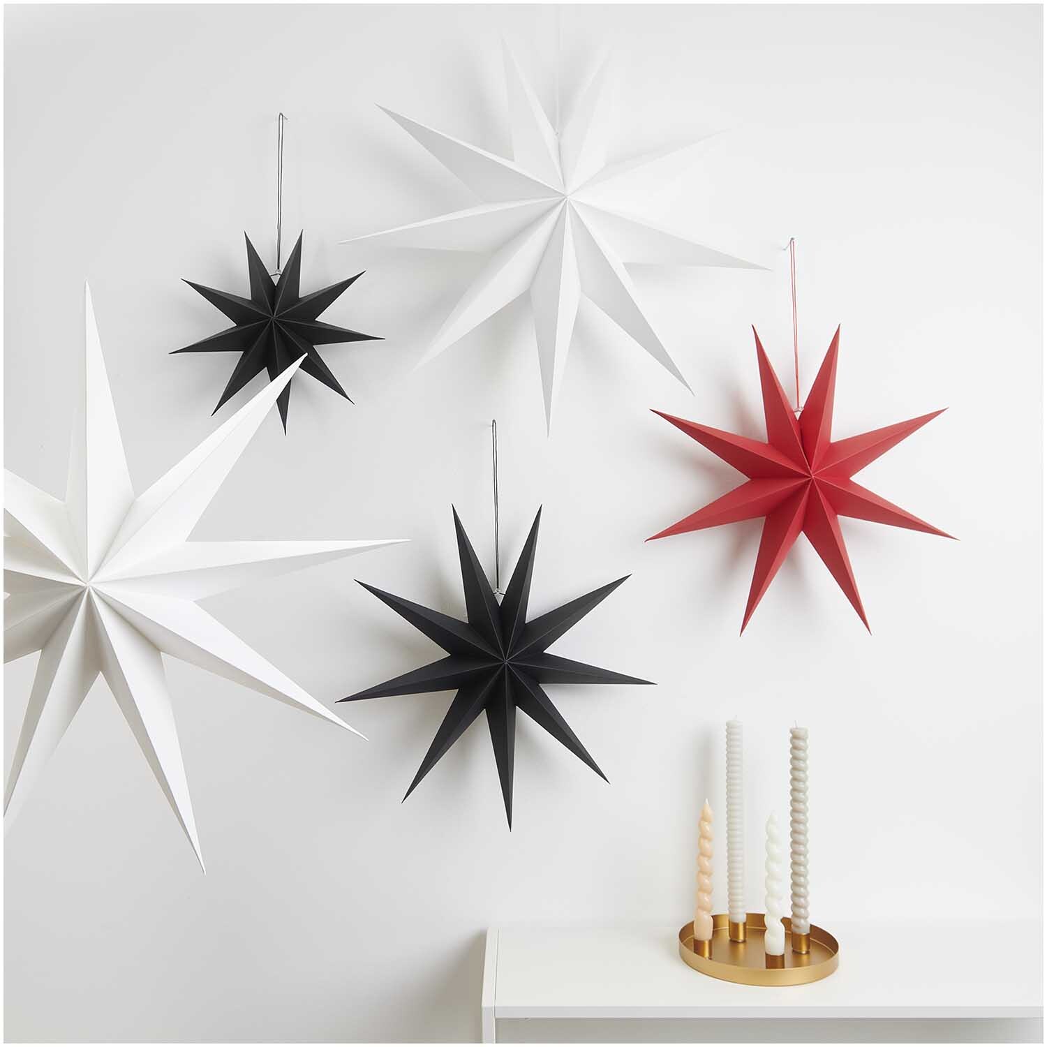 Papierstern auffaltbar 45cm von Rico Design in Weiß, umgeben von weiteren Sternen in Schwarz und Rot an einer Wand.