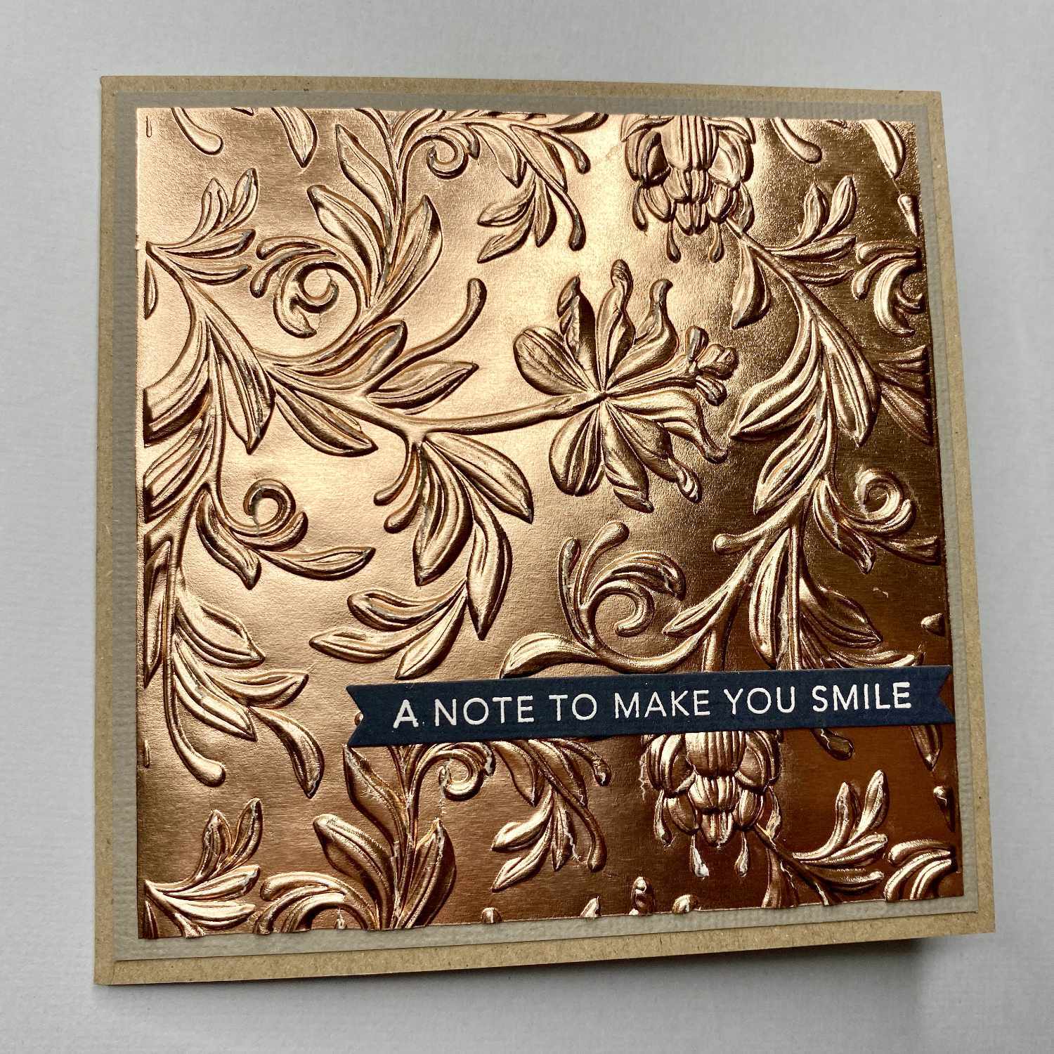 3D Textured Fades Embossing Folder Botanical: Prächtiges, florales Muster in Kupferoptik auf einer Grußkarte mit dem Text "A Note to Make You Smile".