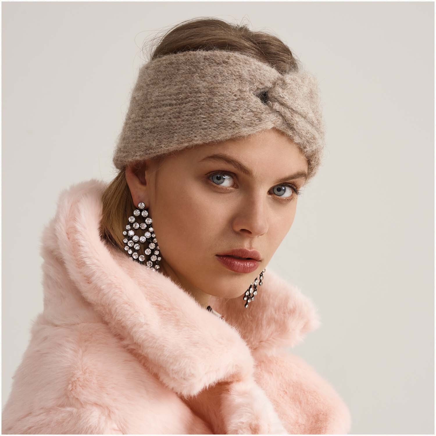 Luxury Volumi Stirnband in Beige, getragen von Frau in rosa Mantel. Elegante Ohrringe ergänzen den Look.