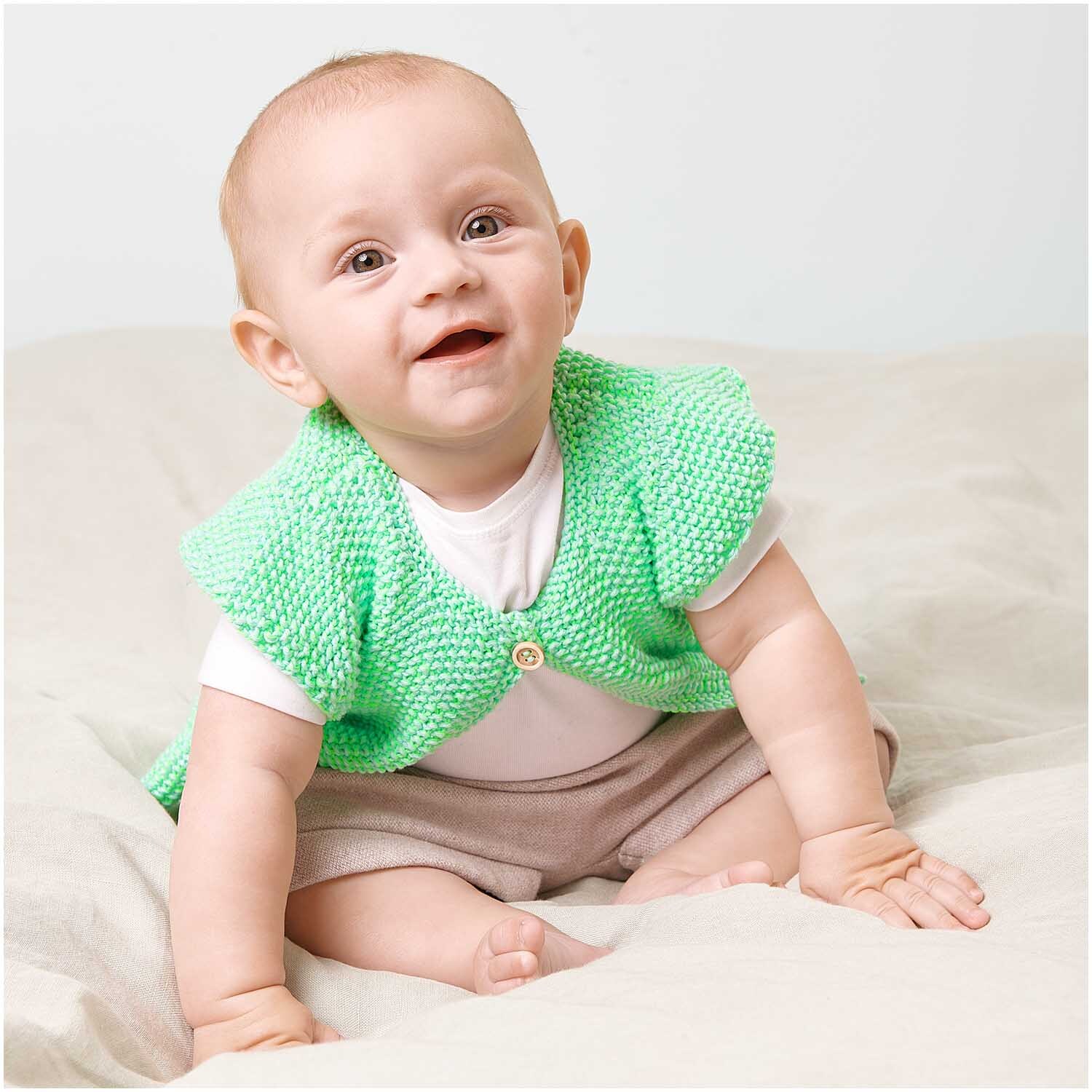 Creative Make It Neon: Ein lächelndes Baby trägt eine grüne Strickweste von Rico Design auf einem Bett.