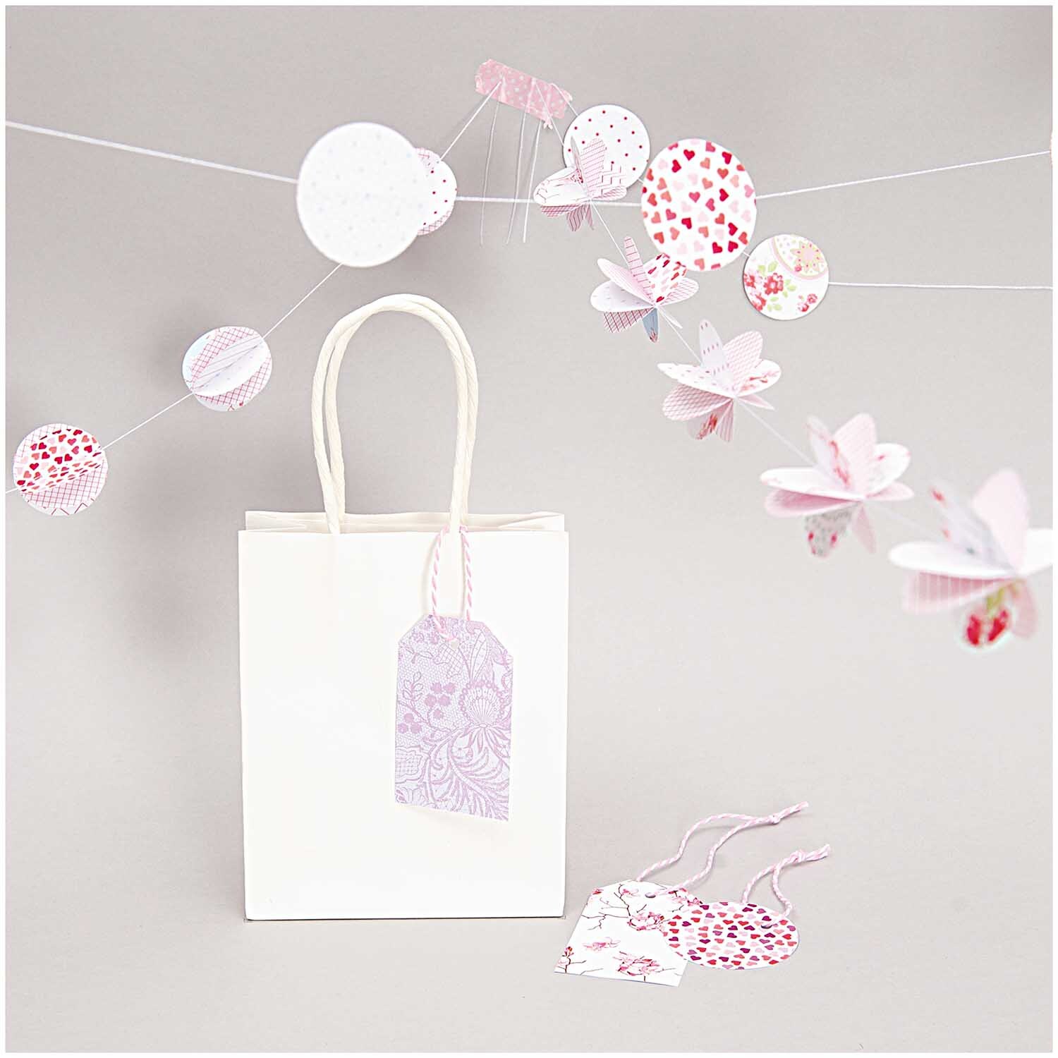 Paper Poetry Baumwollgarn rosa-weiß 15m von Rico Design, dekoriert mit zarten Papierblumen und Geschenktüte.