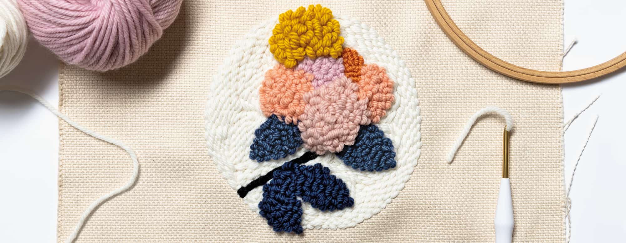 Ein Blumenmuster ist mit der Punch Needle auf Stoff gestickt. Punch Needle, Stickrahmen und Wollknäuel liegen daneben. 