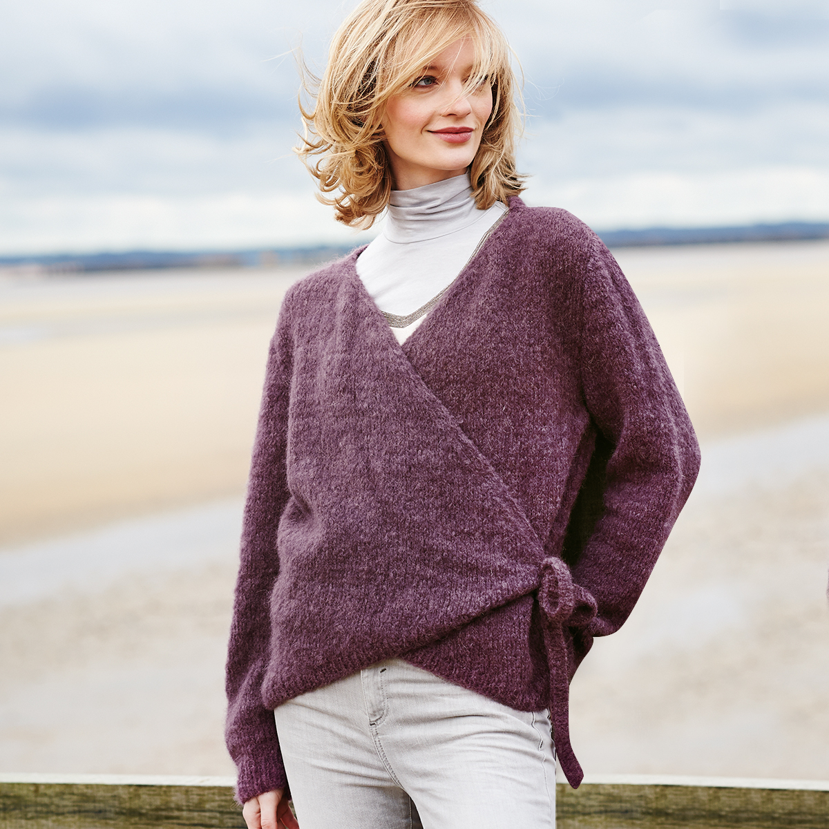 Strickanleitung Wickeljacke aus Rico Luxury Volumi: Frau am Strand trägt lila Wickeljacke über grauem Rollkragenpullover.