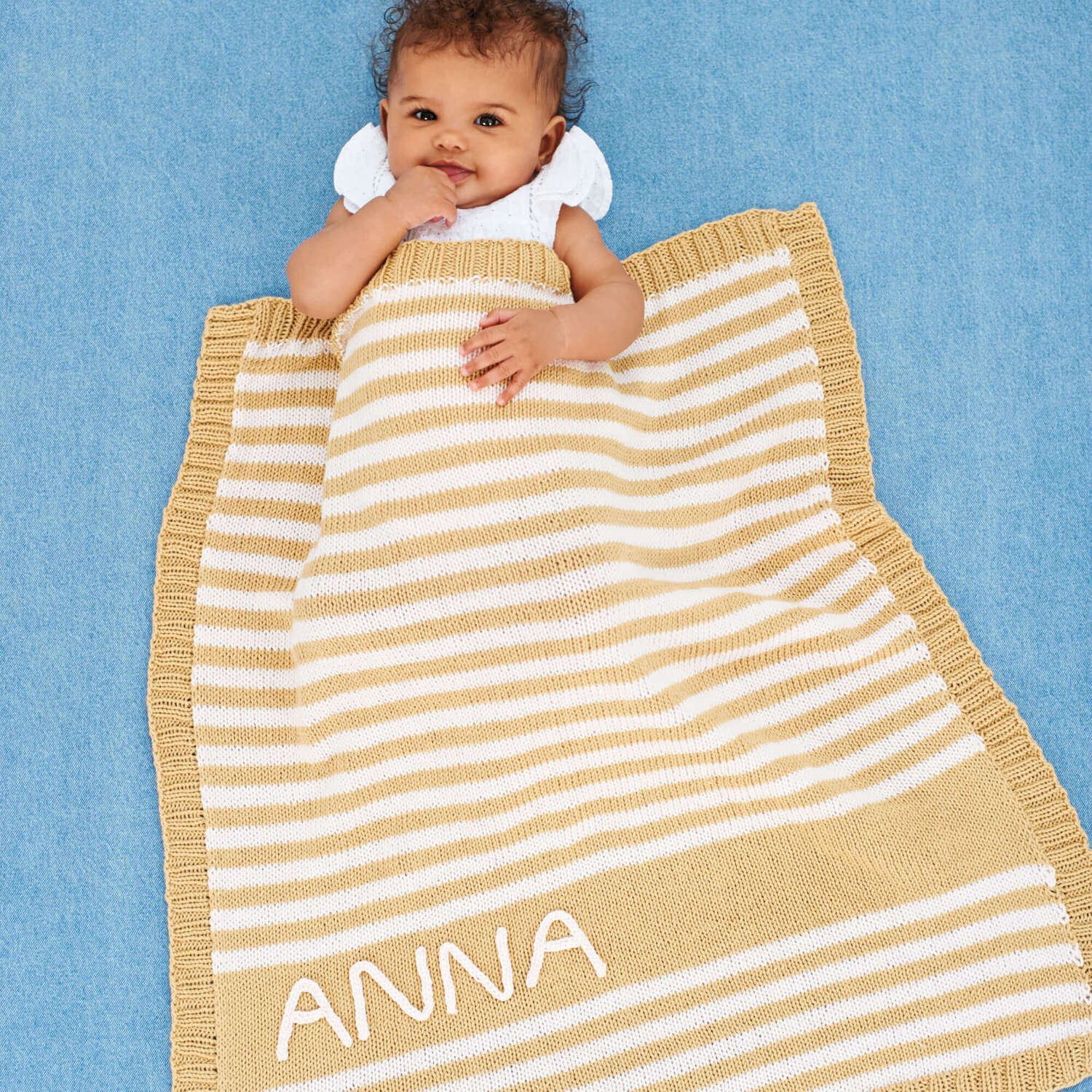 Strickanleitung Babydecke mit Streifen: Baby liegt auf blauer Decke, eingewickelt in gestreifte Decke mit Namen "ANNA".