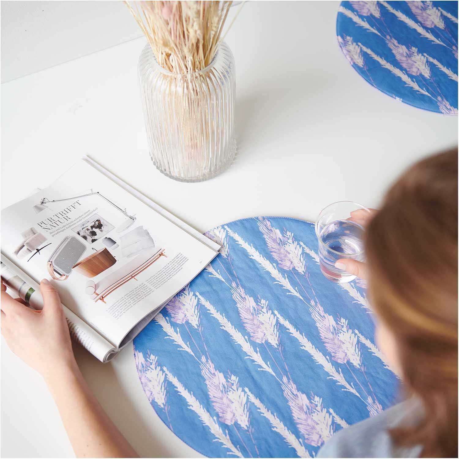 Meterware Druckstoff Pampasgras azur: Tischset mit Pampasgras-Muster in Blau, neben Vase und Magazin.