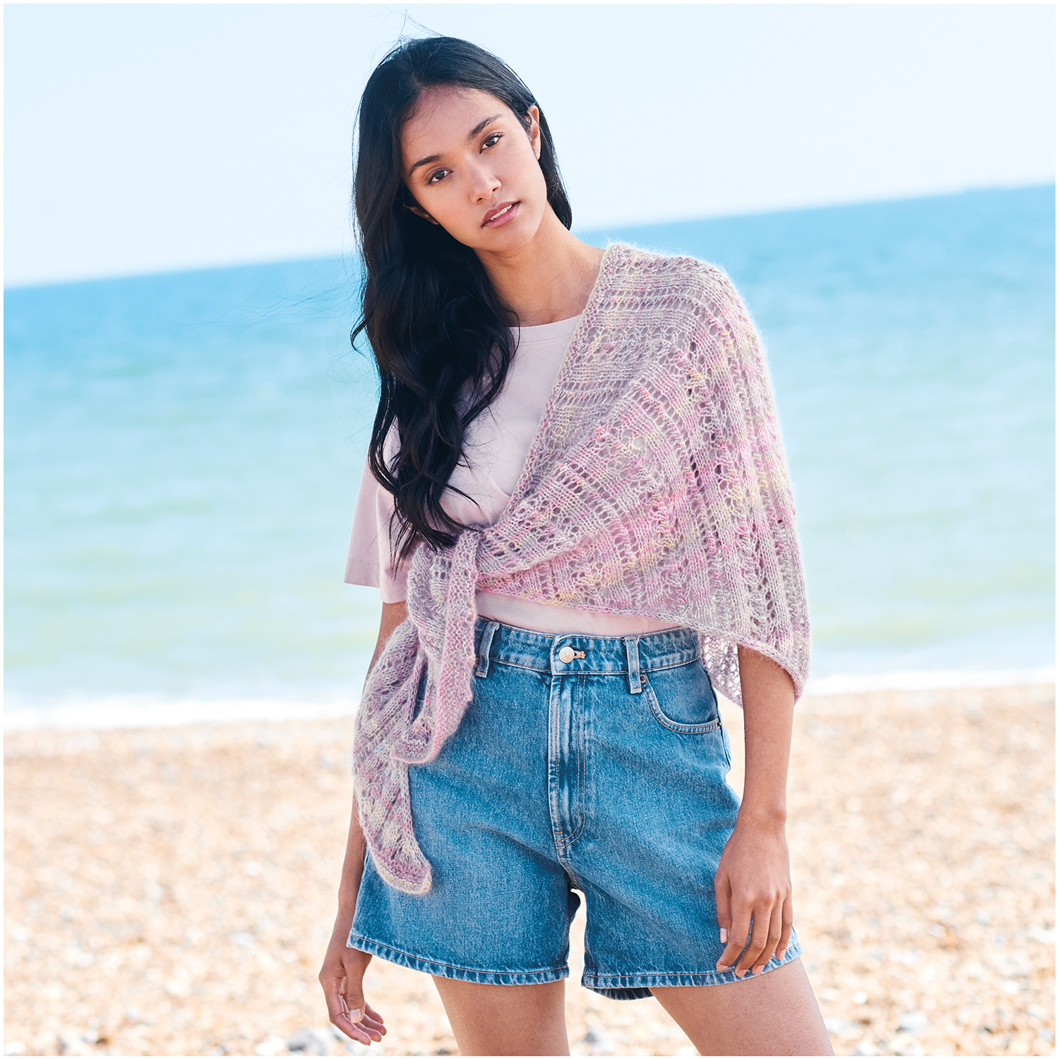 Strickset Tuch Modell 13 aus Lovewool Nr. 18: Frau am Strand mit rosa Tuch und Jeansshorts.
