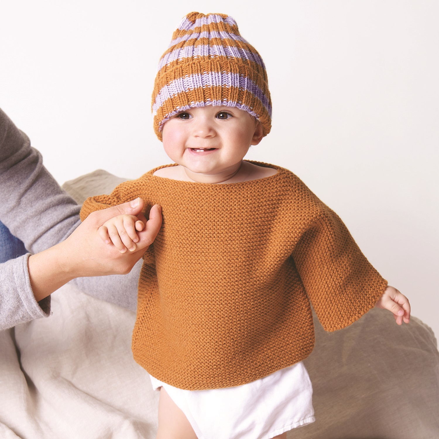 Strickanleitung Pullover & Mütze aus Baby Classic dk: Baby trägt gestrickten Pullover und gestreifte Mütze, gehalten von einer Hand.