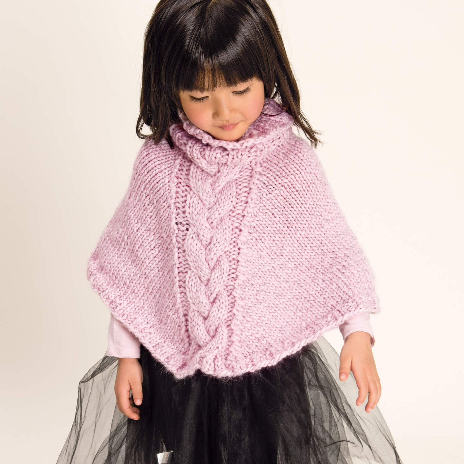 Strickanleitung Kinderponcho aus Glühwürmchen Garn: Mädchen trägt rosa Poncho mit Zopfmuster, kombiniert mit schwarzem Rock.