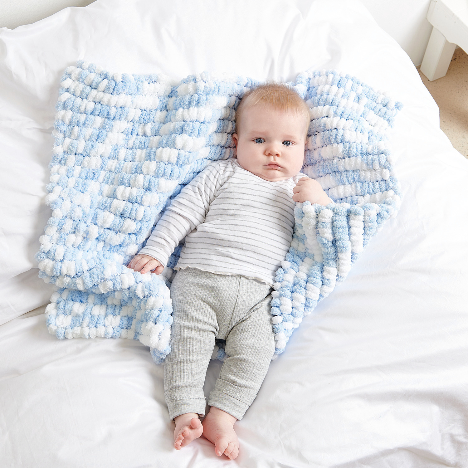 Creative Pompon Print: Baby in weicher, babyblauer Decke von Rico Design, 200g, auf weißem Bett.