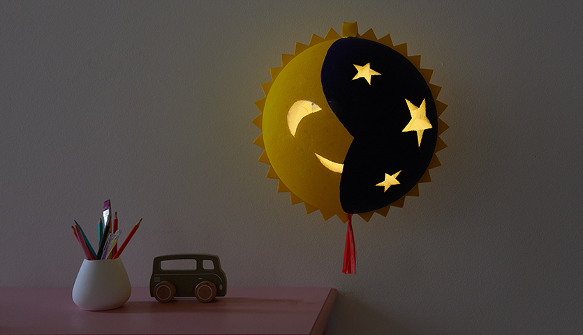 Bastelanleitung Sonne Mond Laterne: Eine leuchtende Laterne mit Sonnen- und Mondmotiv, dekoriert mit Sternen, auf einem Tisch mit Stiften und Spielzeugauto.