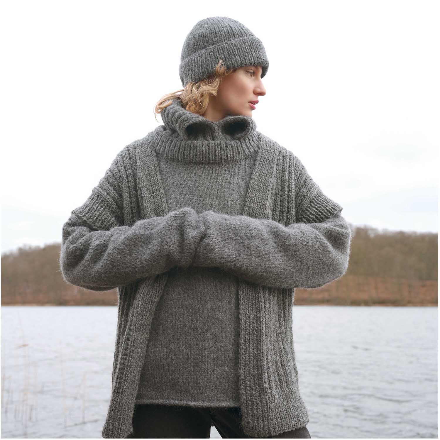 Strickset Pullover Modell 17: Frau in grauem Strickpullover und Mütze vor einem See im Winter.