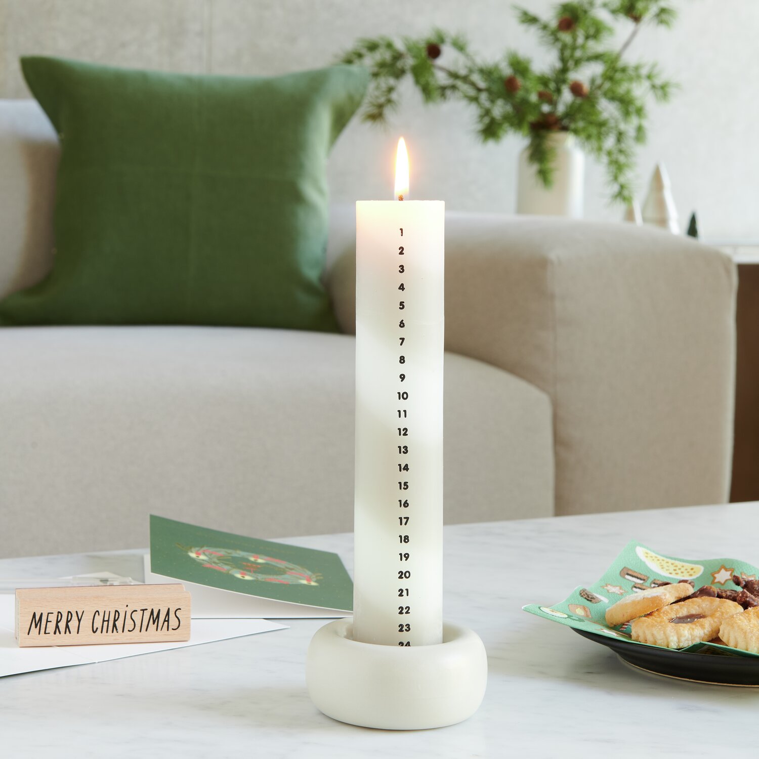 Adventskalender Kerze gestreift, Beige-Weiß, Ø4x20cm, brennend auf Tisch, im Hintergrund Sofa.