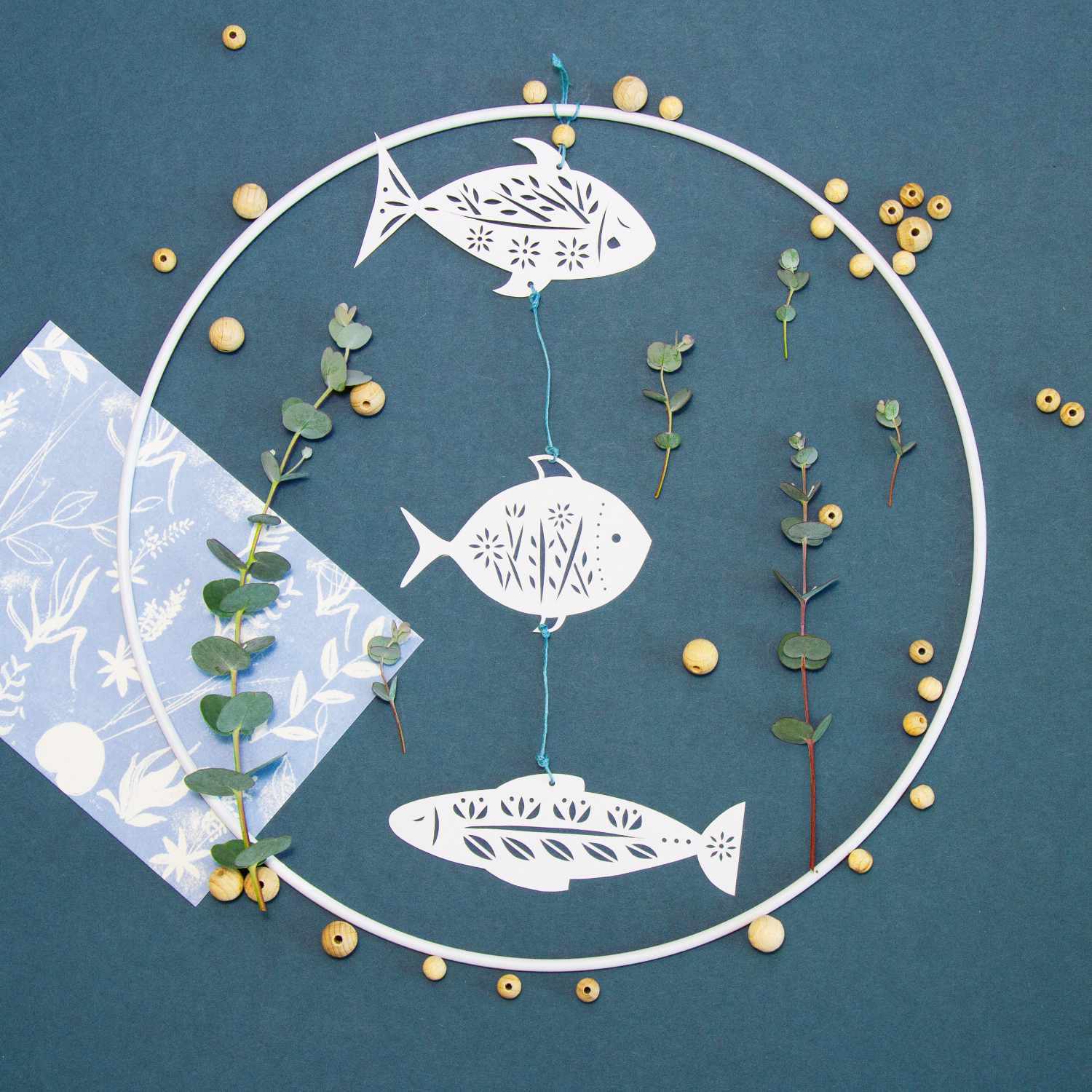 Bastelanleitung Papercut Fische: Drei filigrane Papierfische in einem Kreis, umgeben von Zweigen und Perlen auf blauem Hintergrund.