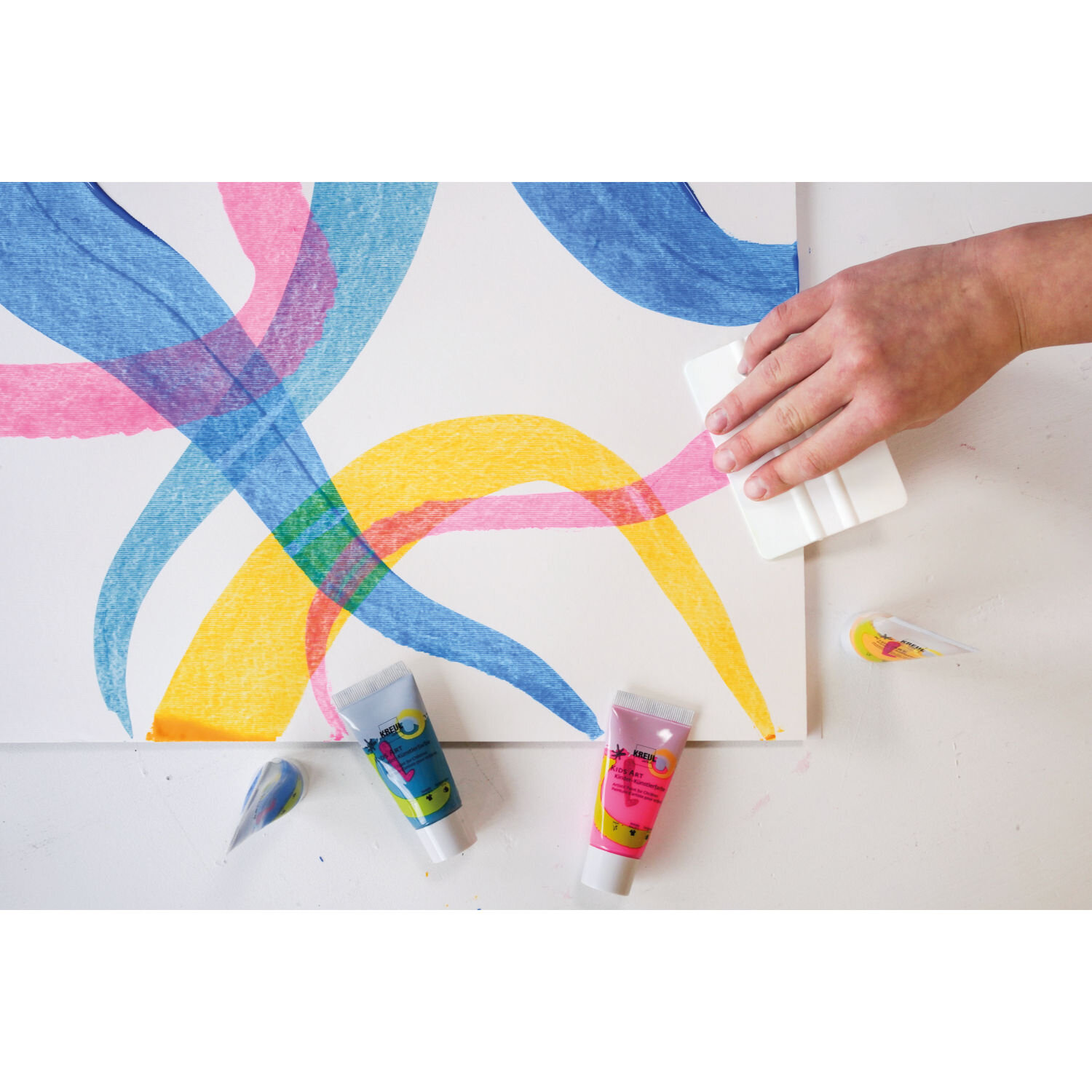 Squeegee Art Set von KREUL mit Spachtel und Farbtuben auf weißer Leinwand, auf der schwungvolle bunte Farbflächen kreativ verwischt werden