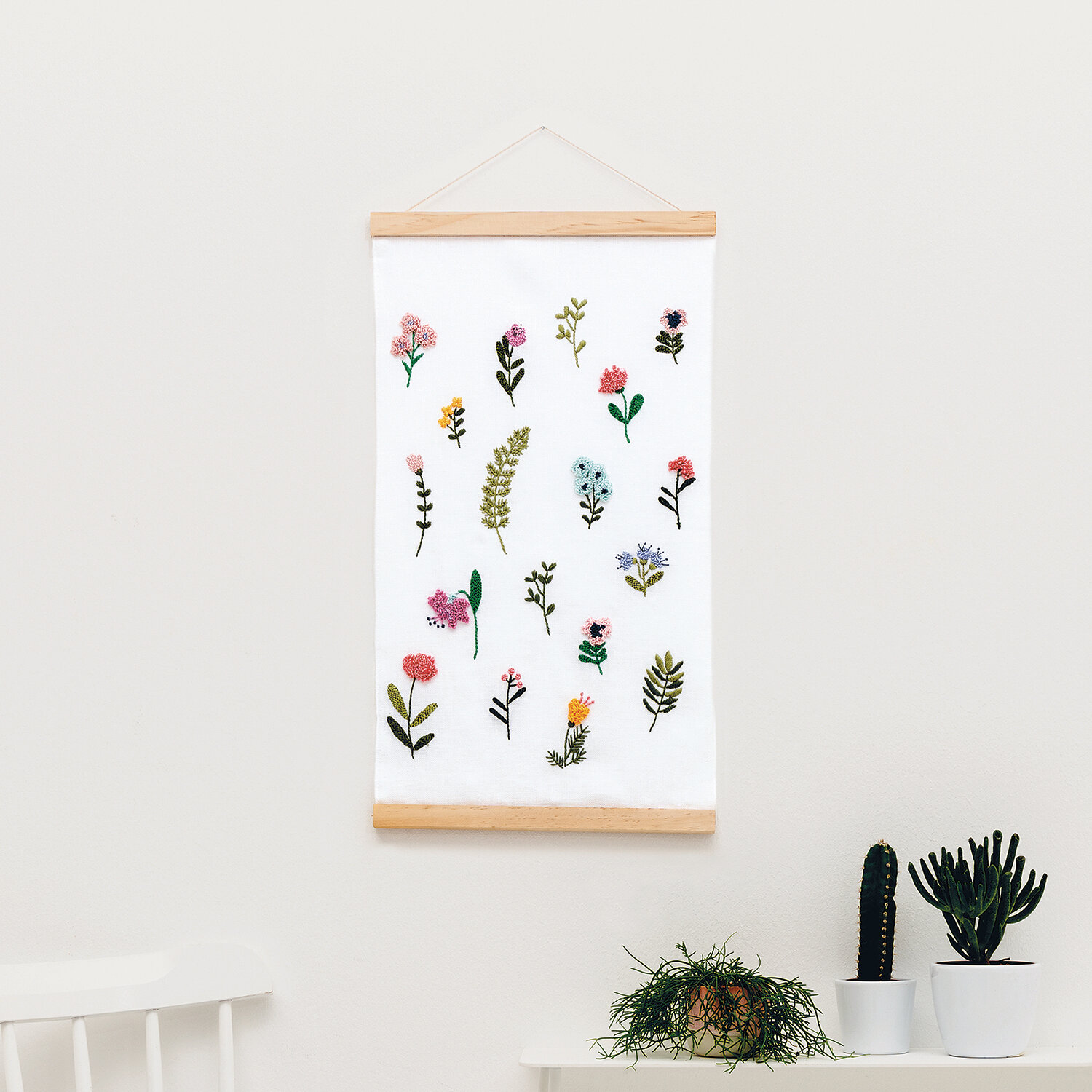 Anleitung Punch Needle Wandbehang mit Blumenwiese zeigt gestickte bunte Blüten auf hellen Stoffbahnen, dekorativ über einem weißen Stuhl und Pflanzen platziert