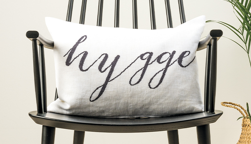 Stickanleitung Hygge Kissen: Weißes Kissen mit schwarzem Schriftzug "hygge" auf einem schwarzen Stuhl.