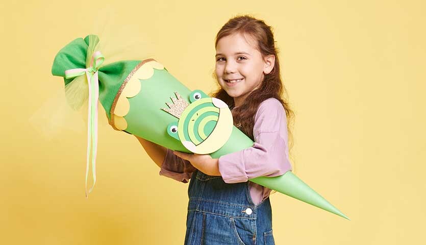Bastelanleitung Schultüte Frosch: Mädchen hält grüne Schultüte mit Froschmotiv vor gelbem Hintergrund.