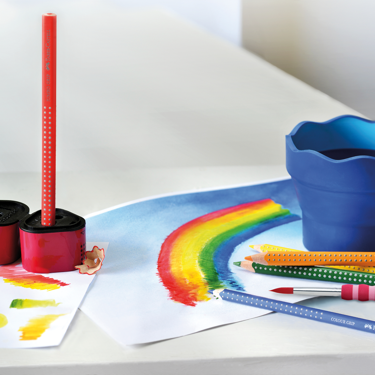 Jumbo Grip Farbstift von Faber Castell auf einem Tisch mit buntem Regenbogenbild und Malzubehör.