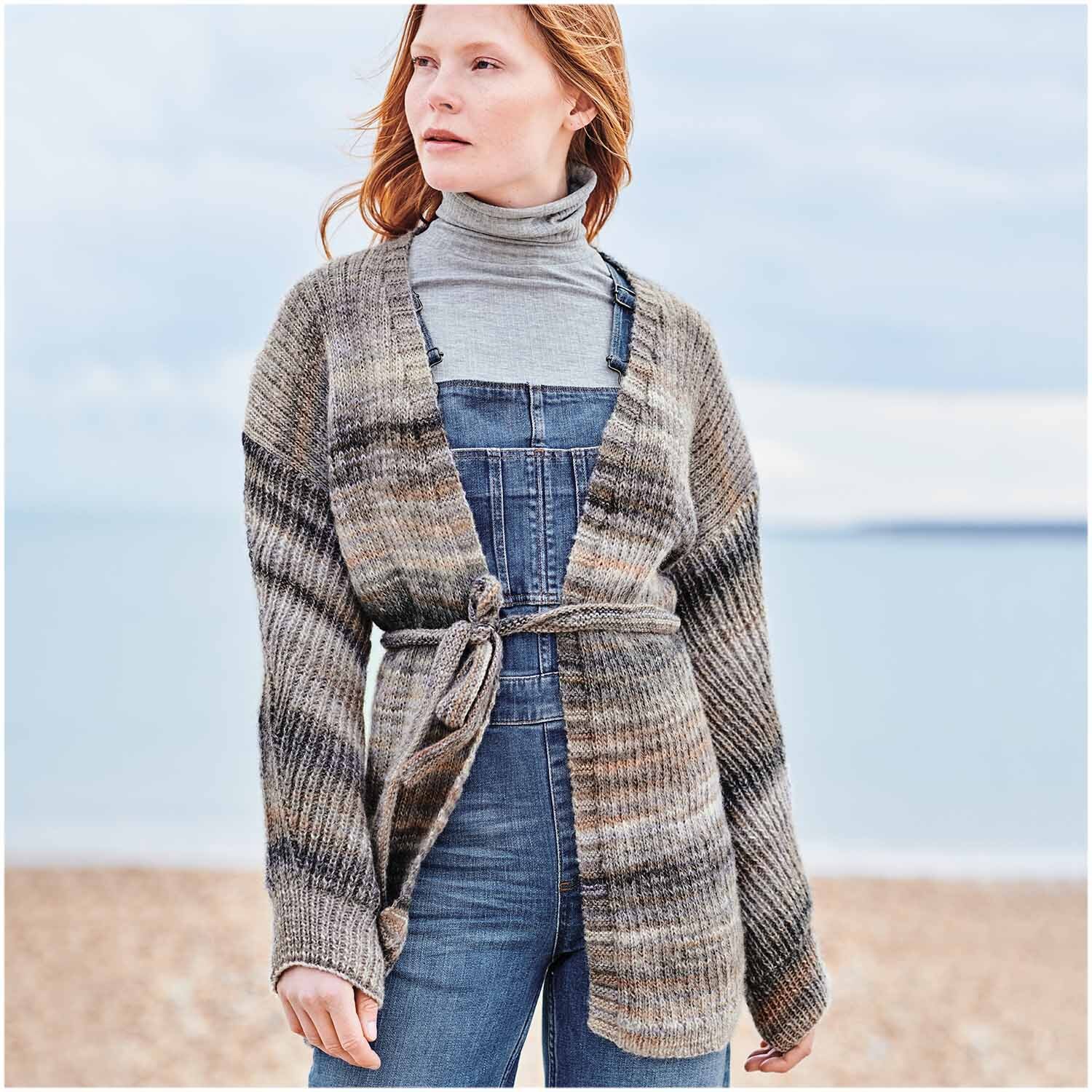 Creative Melange Garzato aran Wonderball: Frau am Strand in gestrickter Strickjacke in Naturtönen, kombiniert mit Jeans und Rollkragenpullover.