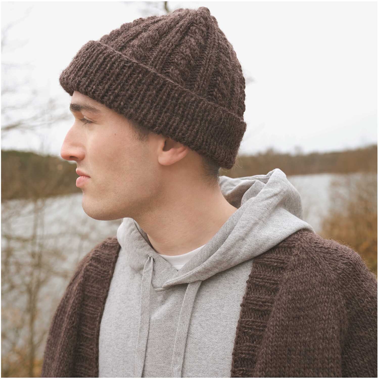 Strickset Mütze Modell 03 aus Luxury Knits Winter Special: Braune Mütze, getragen von einem Mann in grauem Hoodie, vor herbstlicher Landschaft.